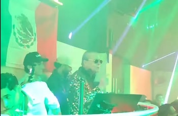 Una persona con gafas de sol y camisa estampada frente a una consola en una discoteca con luces verdes y haces de láser, y una bandera mexicana al fondo