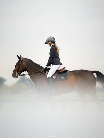 Cande Tinelli, jinete con casco y chaqueta oscura, monta un caballo marrón al galope, con fondo claro y desenfocado