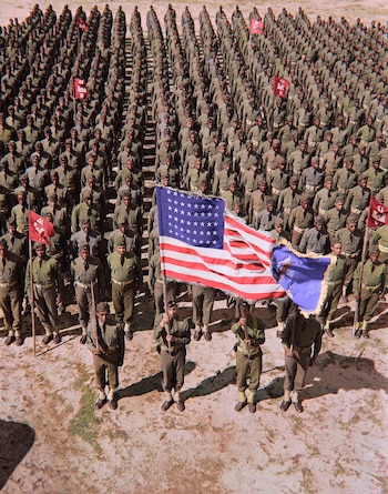 Cientos de soldados afroamericanos en uniformes militares verdes oliva forman en terreno arenoso. Un grupo en primera fila sostiene la bandera de EE. UU. y otras banderas rojas