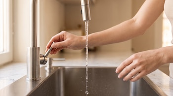 Primer plano de manos de persona abriendo un grifo de acero inoxidable; agua cae en una pileta metálica sobre mesada de mármol.
