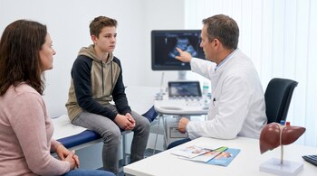 Un médico en bata blanca señala una ecografía en un monitor a un adolescente y una mujer sentados. Sobre el escritorio hay un modelo anatómico de hígado.