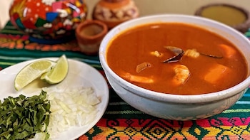 Primer plano de un tazón de sopa de camarones anaranjada rojiza, servida con gajos de lima, cebolla picada y cilantro en un plato blanco