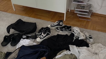 Alfombra clara con un bolso negro, sandalias, tres pares de zapatillas, pantalón de mezclilla, sudadera negra y ropa blanca y verde.