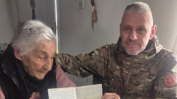Una mujer mayor con cabello blanco y un hombre en uniforme militar camuflado miran una carta escrita a mano sobre una mesa