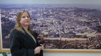 Retrato de primer plano de Dana Erlich, una mujer de cabello rubio, ojos azules y piel clara, usando chaqueta oscura y joyas doradas, con fondo blanco y rejilla