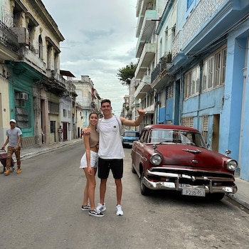 Un hombre y una mujer jóvenes de pie en una calle empedrada, sonríen. Un automóvil clásico rojo estacionado a la derecha. Edificios de varios colores bordean la calle