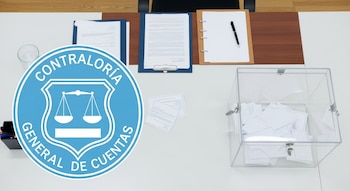 Símbolo de la Contraloría General de Cuentas, documentos en carpetas, vaso de agua, una urna transparente con boletas y bolígrafos sobre una mesa de despacho.