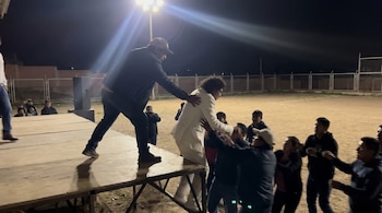 Un hombre de traje blanco es empujado de un escenario por otro hombre con gorra. Varias personas abajo intentan ayudar en un evento nocturno.