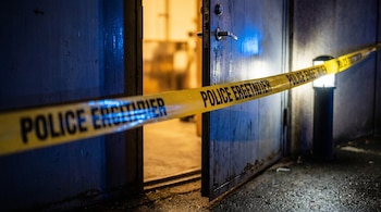 Una cinta policial amarilla con letras negras cruza una puerta de metal azul oscuro semiabierta, con luz cálida en el interior y un reflejo azul de sirena en la puerta.