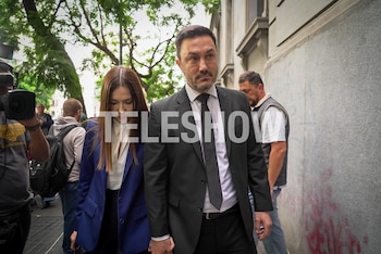 Un hombre de traje oscuro y una mujer de blazer azul y camisa blanca caminan con expresión seria, tomados de la mano, con gente y un camarógrafo detrás