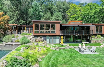 Casa de dos pisos con fachadas de madera y grandes ventanales, un patio con césped verde, sillas de exterior y piscina al costado