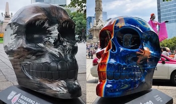 Dos esculturas de cráneos decoradas con pintura sobre pedestales en una avenida pública con monumentos urbanos al fondo