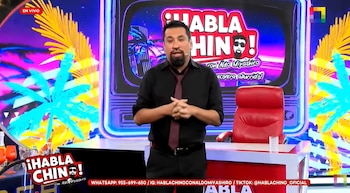 Hombre con barba y camisa oscura de pie en un set de televisión. Logo '¡Habla Chino!', fondo de neón con palmeras, silla roja, escritorio y texto 'EN VIVO'