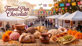 Mesa con conchas, pan de muerto, tamales, flores de cempasúchil y jarros con chocolate, frente a una plaza con carpas y banderas de papel picado.