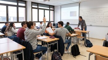 Salón de clase con un profesor de pie frente a alumnos sentados en pupitres, algunos con mochilas. Una estudiante levanta la mano.