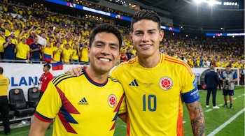James Rodríguez con camiseta amarilla de Colombia y un aficionado sonríen juntos en un campo de fútbol con gradas llenas de gente al fondo.