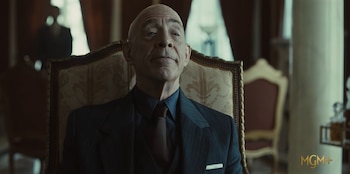 Hombre calvo vestido con traje, camisa oscura y corbata, sentado en un sillón. Fondo difuso con otra persona y decoración. Logotipo de MGM+