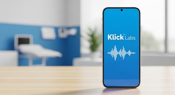Teléfono inteligente con logo Klick Labs y ondas de voz en pantalla azul, sobre mesa de madera clara, con consultorio médico difuminado al fondo.