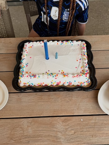 Pastel rectangular blanco con glaseado y grageas de colores en los bordes, dos velas azules, bandeja negra, mesa de madera, dos platos, persona con camiseta deportiva