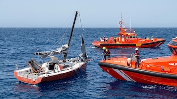 Dos embarcaciones naranjas de Salvamento Marítimo junto a un velero rojo con velas recogidas sobre el agua.