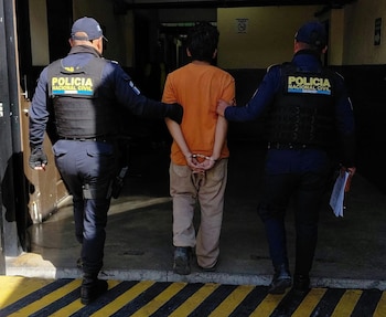 Dos agentes con chalecos de la Policía Nacional Civil sujetan del brazo a un hombre esposado que viste camiseta naranja