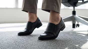 Pies con zapatos negros que elevan los talones sobre el suelo alfombrado junto a una silla de oficina.