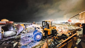 Una cargadora frontal amarilla en primer plano, rodeada de escombros de madera, metal y lona en una playa por la noche. Al fondo, el océano y la silueta de edificios iluminados