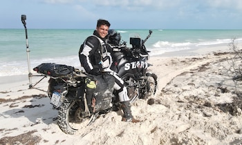Hombre sonriente en equipo de motociclismo blanco y negro sentado en una motocicleta de aventura en una playa de arena con el mar turquesa de fondo