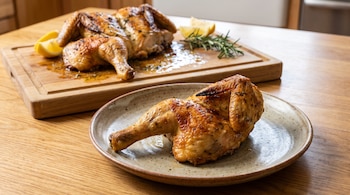 Un cuarto de pollo asado en un plato beige, el resto del pollo asado en una tabla de madera con rodajas de limón y ramitas de romero.