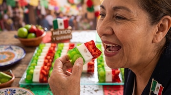 Una mujer sostiene una gelatina verde, blanca y roja cerca de su boca frente a una bandeja con postres y adornos mexicanos.