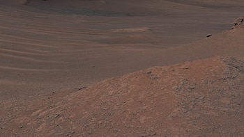 Vasto paisaje árido de arena rojiza y marrón con dunas suaves y ondulaciones. En primer plano, terreno rocoso y fragmentado