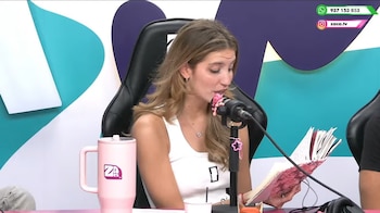 Una joven de cabello claro sostiene y lee un cuaderno rosado frente a un micrófono de mesa junto a un vaso térmico rosa