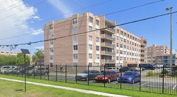 Edificio residencial de ladrillo de varios pisos con balcones y un aparcamiento exterior lleno de coches y furgonetas, rodeado por una valla negra