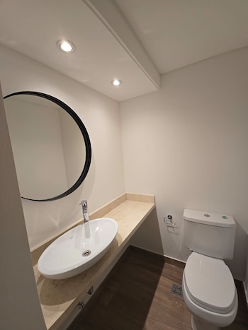 Baño moderno con paredes blancas, lavabo ovalado sobre encimera de mármol beige, espejo redondo con marco negro, inodoro blanco y suelo de madera oscura
