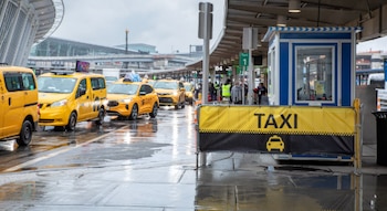 Fila de taxis amarillos estacionados en una zona de espera de aeropuerto con el suelo mojado. Se observa una caseta de información y un cartel de "TAXI"