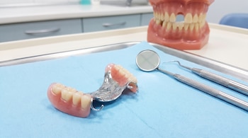 Una prótesis dental parcial removible de metal y resina sobre un paño azul, junto a un espejo y dos exploradores dentales; al fondo, un modelo dental.