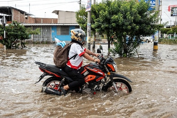 Un motociclista con casco y mochila avanza sobre una motocicleta que cruza una calle urbana cubierta por agua marrón