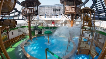 Imagen de un parque acuático con piscinas, toboganes marrones, estructuras de madera, red de escalada y abundante agua salpicando