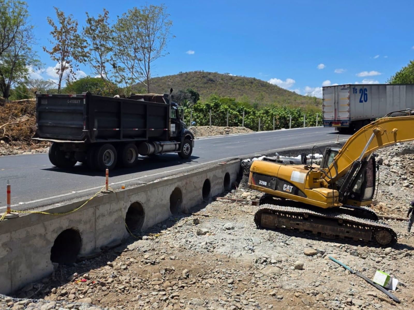 Una excavadora Caterpillar realiza trabajos de infraestructura junto a una estructura de drenaje de concreto mientras circulan camiones de carga por la carretera. (CIV Guatemala)