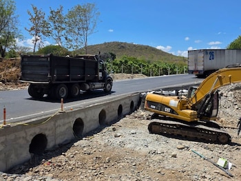 Una excavadora amarilla Caterpillar junto a un muro de concreto con alcantarillas circulares y un camión negro que transita por la vía