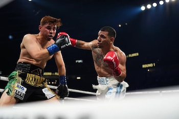 Dos boxeadores en el ring; uno a la izquierda con shorts negros y verdes y guantes azules, el otro a la derecha con shorts blancos y guantes rojos, lanzando un puñetazo