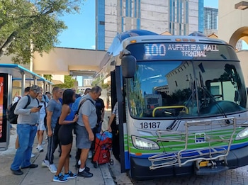 Grupo de personas haciendo fila para abordar un autobús público en una parada urbana soleada. El autobús muestra '100 Aventura Mall Terminal'