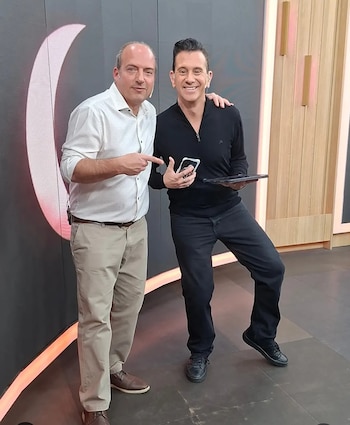 Dos hombres de pie en un estudio. El de la izquierda señala al de la derecha, quien sostiene un teléfono móvil y una tableta