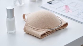 Un sostén beige con varilla metálica visible junto a un envase de vidrio esmerilado y un portapapeles con un lazo rosa.