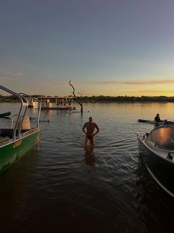 Hombre en bañador sale del agua al atardecer, entre dos barcos. Una balsa flotante con personas y árboles distantes bajo un cielo anaranjado