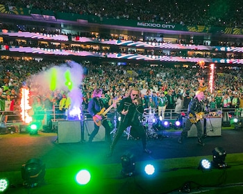 Tres músicos de rock en escenario con luces, fuego y humo. Público en gradas de un estadio con cartel MEXICO CITY y banderas