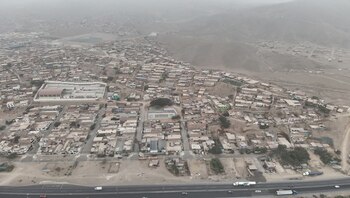 Vista aérea de un asentamiento urbano denso en laderas áridas con casas de ladrillo y techos claros, una carretera principal y edificios más grandes