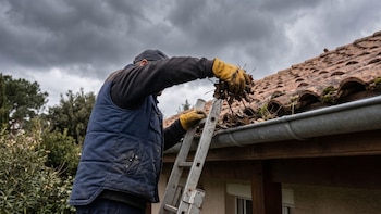 Un hombre con guantes y chaleco sobre una escalera retira hojas secas del tejado y de la canaleta de una casa bajo un cielo nublado