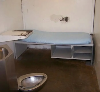 Una celda de prisión pequeña y austera con una cama, un lavabo y un inodoro metálicos. Las paredes son claras y con algunas manchas