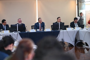 Cinco hombres con traje están sentados en una mesa larga con monitores, micrófonos y carteles de identificación